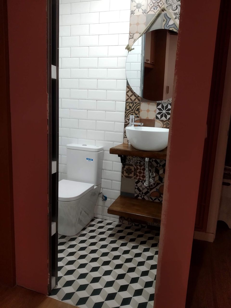 Aseo terminado en el hueco de un antiguo armario: azulejo metro blanco, pared de hidráulico de colores, suelo geométrico, inodoro y lavabo sobre balda de madera, visto desde la puerta