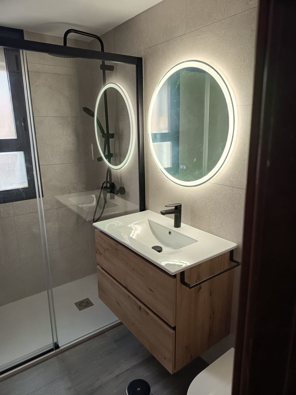 Detalle del baño terminado: espejo redondo retroiluminado, mueble suspendido de roble con lavabo blanco, grifería negra y mampara de perfil negro