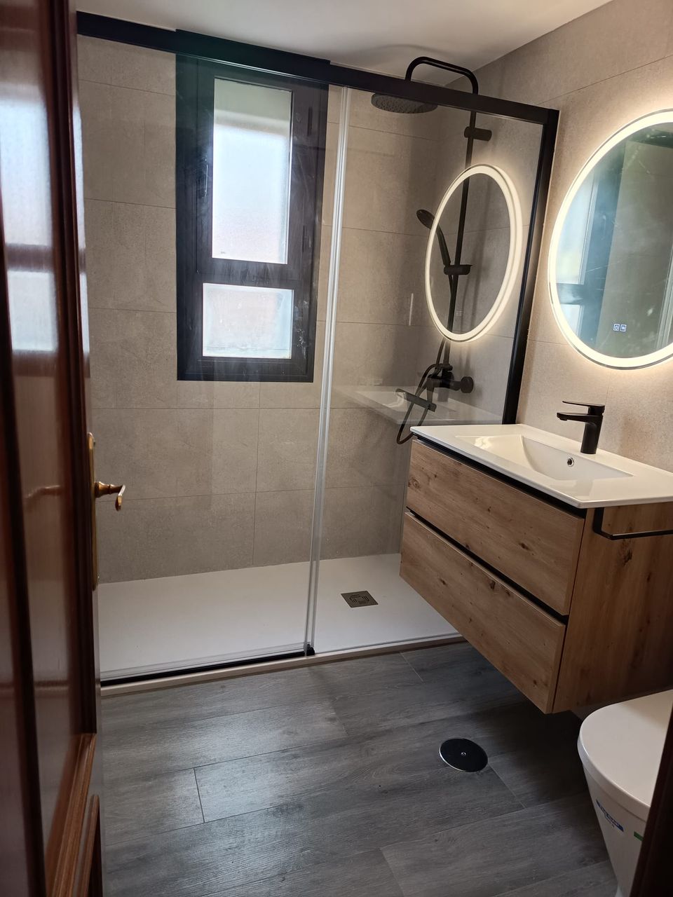 Baño terminado: ducha con mampara de perfil negro, columna de ducha negra, mueble suspendido de roble con lavabo blanco, espejo redondo retroiluminado y suelo porcelánico gris efecto madera