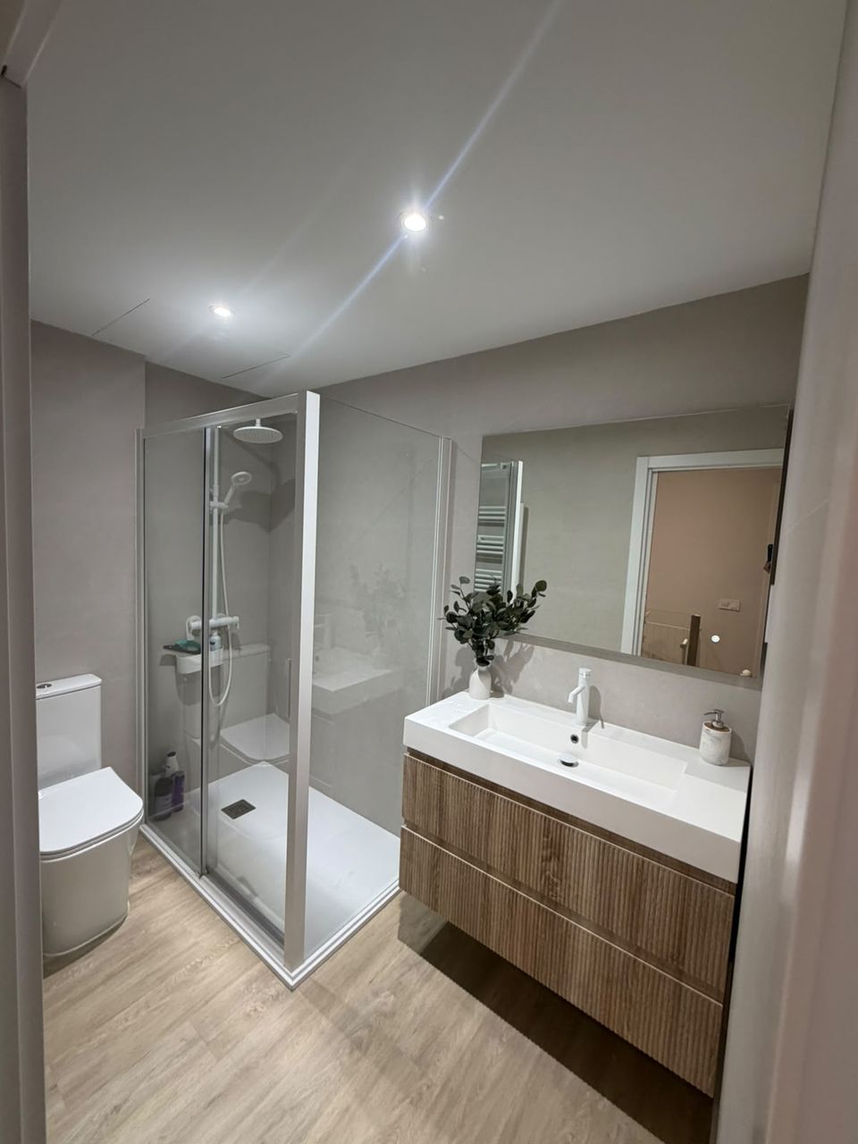 Baño reformado: ducha con mampara de perfil blanco, mueble suspendido de madera estriada con lavabo encastrado, espejo grande y suelo de madera