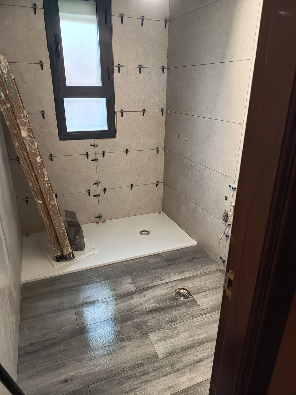 El baño durante la obra: porcelánico gris claro recién colocado con crucetas, plato de ducha blanco y suelo efecto madera gris
