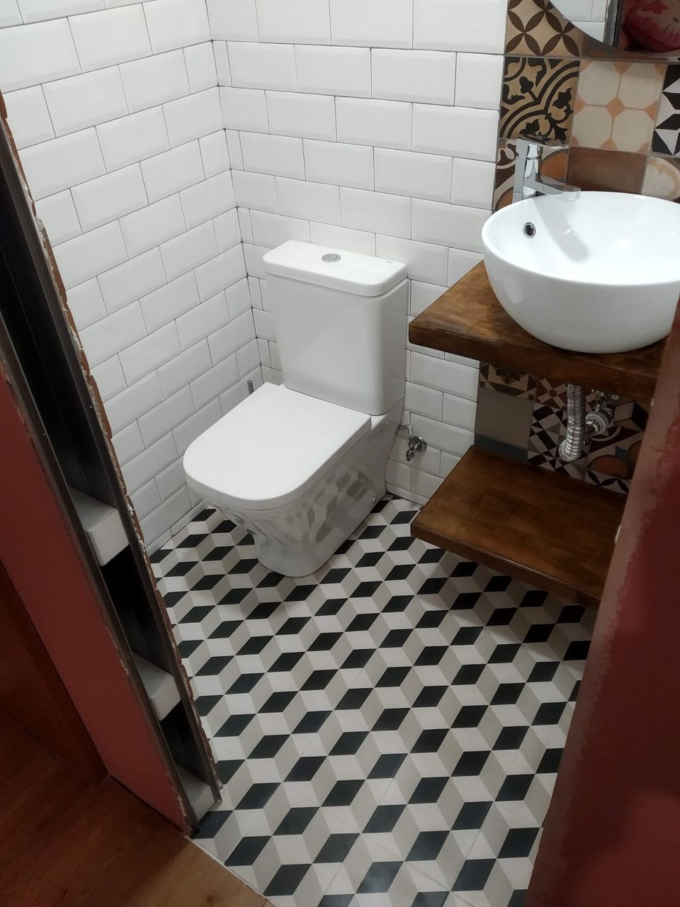 Detalle del aseo: inodoro, lavabo redondo sobre balda de madera, grifería cromada y suelo con dibujo geométrico en blanco, gris y negro