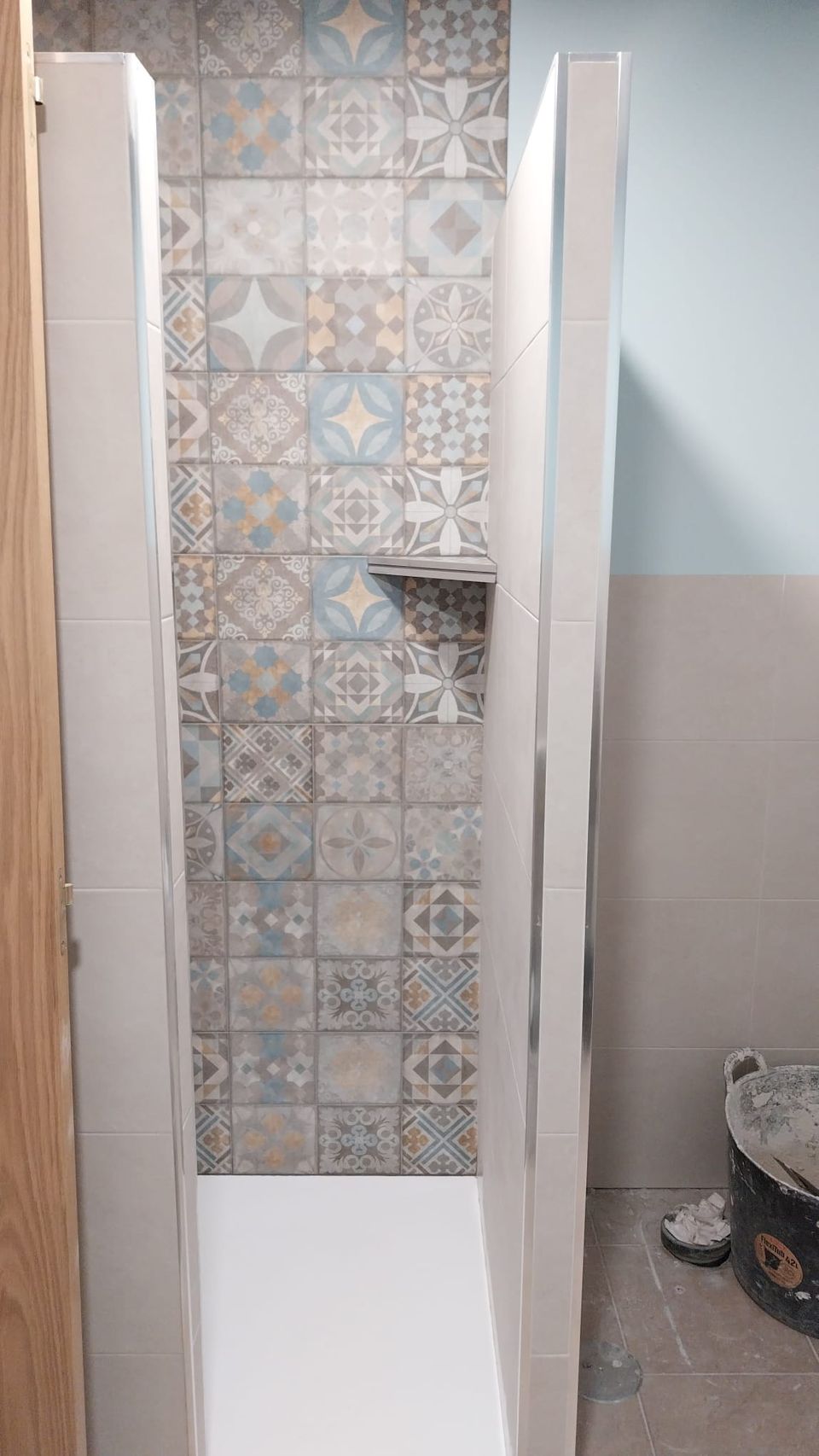 Ducha de obra terminada: fondo de azulejo hidráulico en azules y ocres, repisa de esquina, paredes laterales en porcelánico claro y plato de ducha blanco