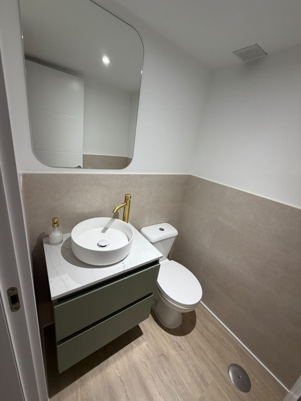 Aseo reformado: mueble suspendido verde salvia con encimera de mármol, lavabo redondo, grifería dorada, espejo de esquinas redondeadas y suelo de madera