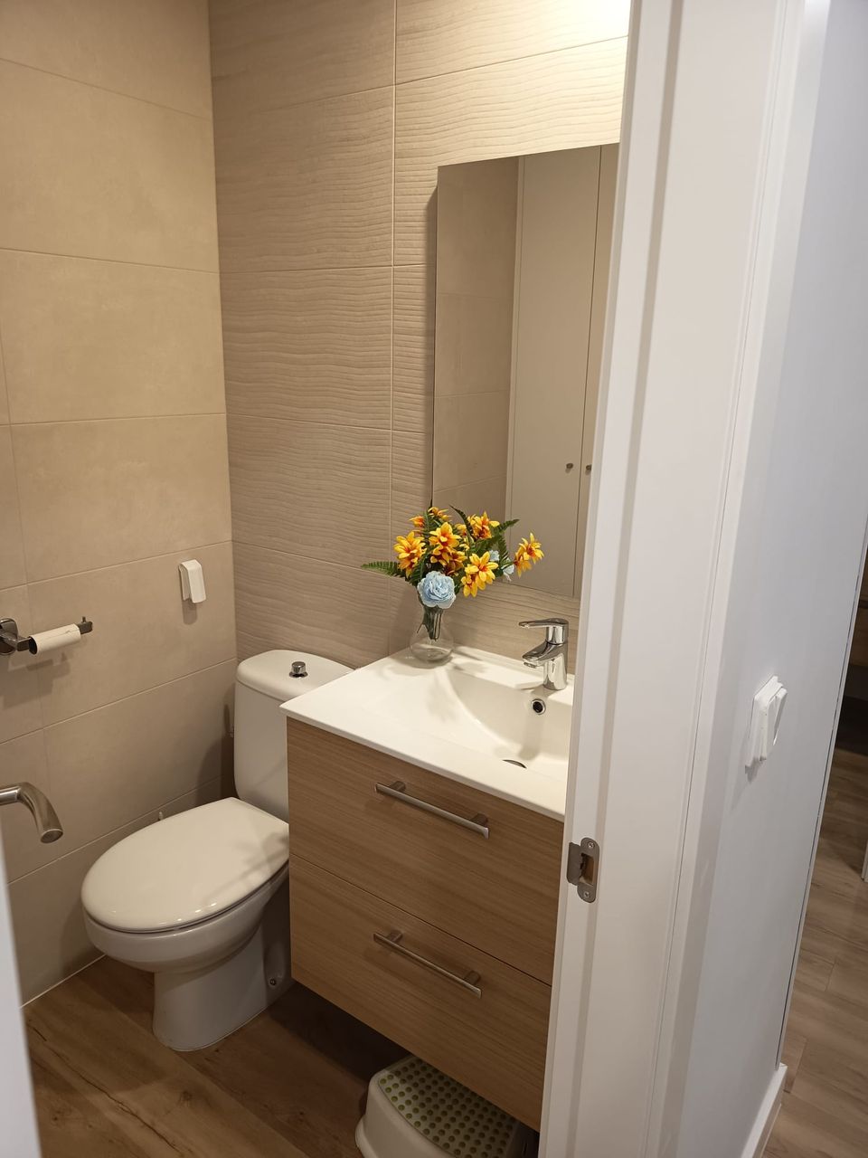 Baño terminado: porcelánico beige en paredes, mueble suspendido de madera clara con lavabo blanco, espejo con armario, inodoro y suelo porcelánico efecto madera