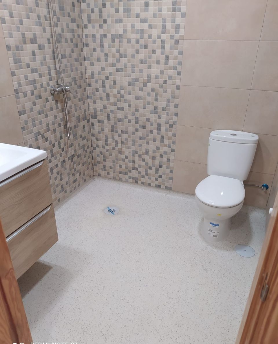 Baño adaptado terminado: ducha abierta con mosaico, suelo continuo antideslizante, inodoro y mueble de lavabo en madera
