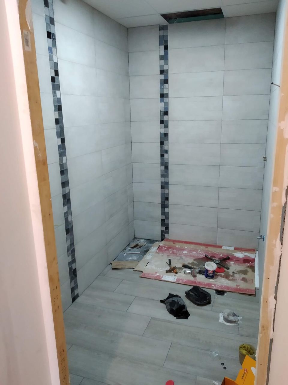 El baño durante la obra: alicatado gris con cenefa vertical de mosaico recién colocado, sin sanitarios