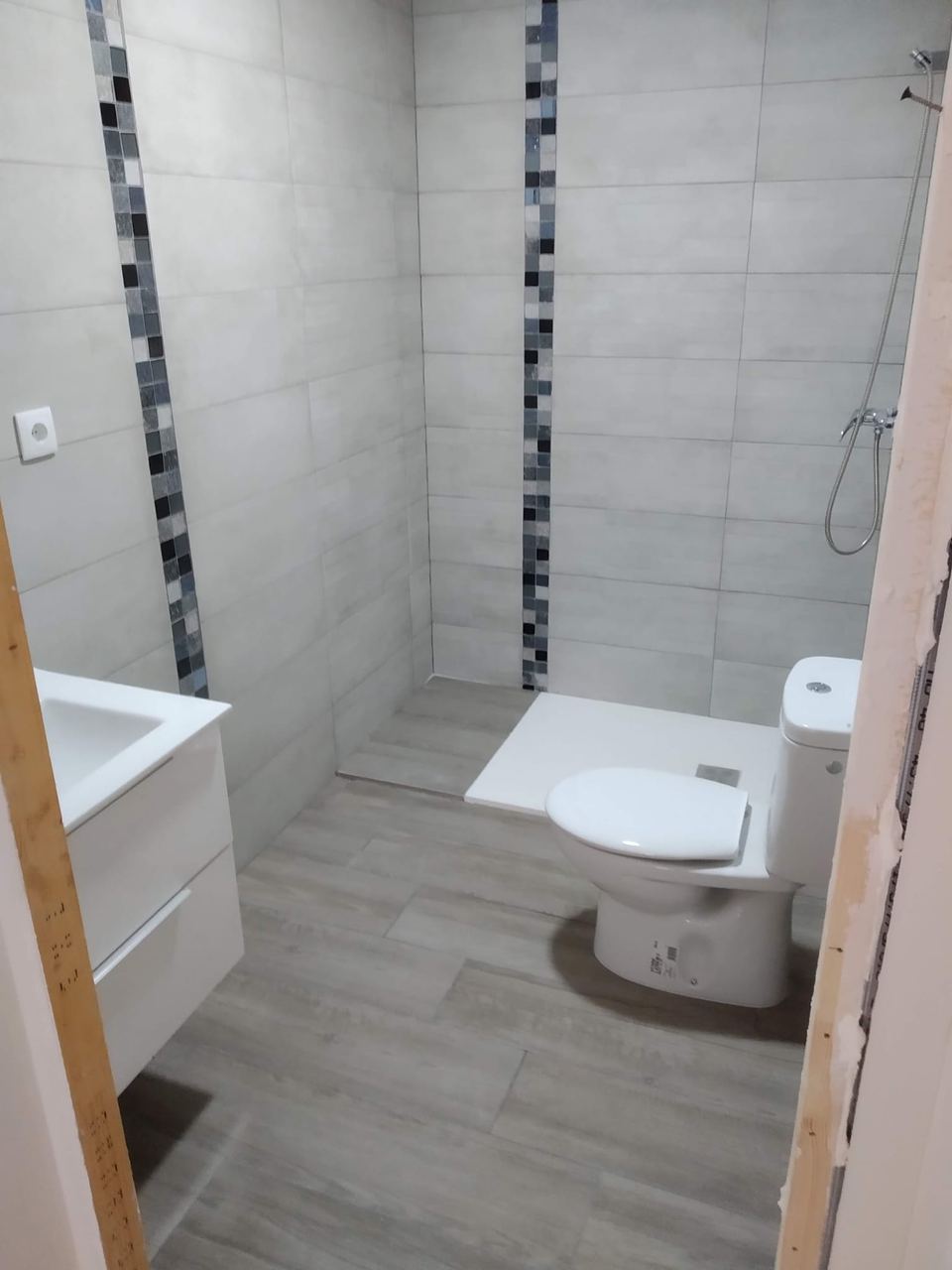 El mismo baño terminado: cenefa vertical de mosaico, plato de ducha, inodoro, mueble suspendido y suelo efecto madera
