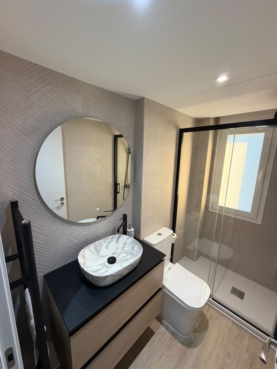 Baño reformado: mueble de madera con encimera negra, lavabo de mármol, grifería negra, espejo redondo, pared con relieve en espiga y ducha con mampara de perfil negro