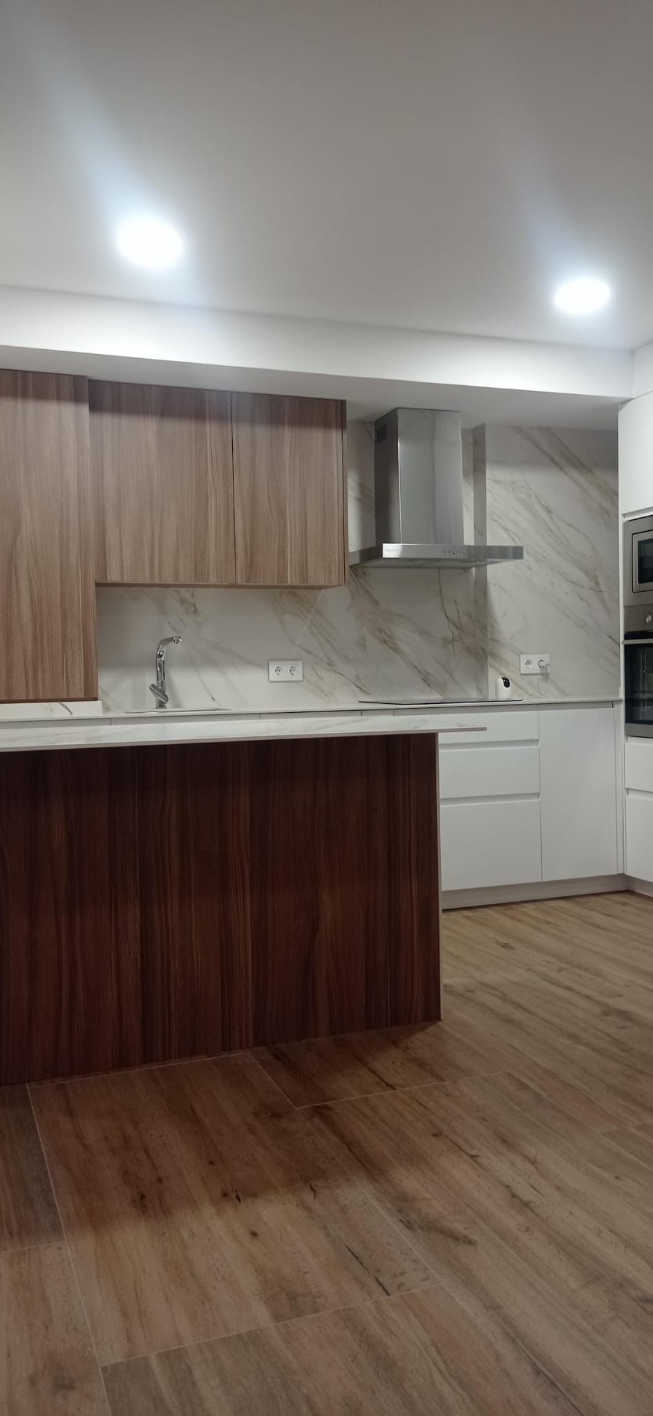 Cocina terminada: frontal porcelánico efecto mármol, muebles altos en madera, bajos en blanco, isla en madera oscura, campana de acero y suelo efecto madera